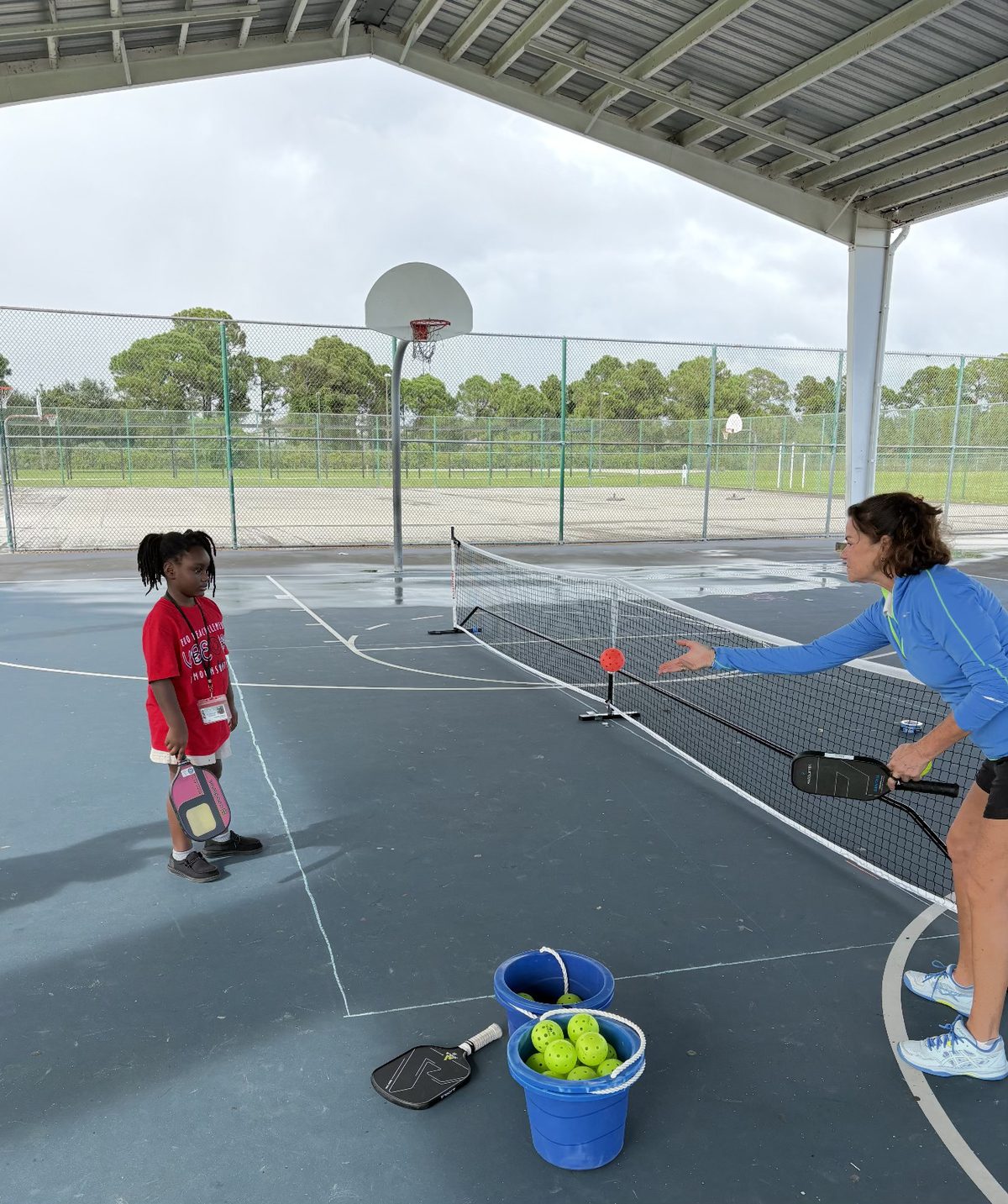 A coach feeding a ball to a young player under a covered court.